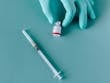 A gloved hand holding a COVID-19 vaccine vial next to a syringe on a teal background.