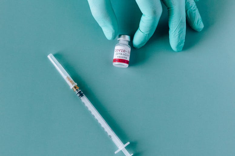 A gloved hand holding a COVID-19 vaccine vial next to a syringe on a teal background.