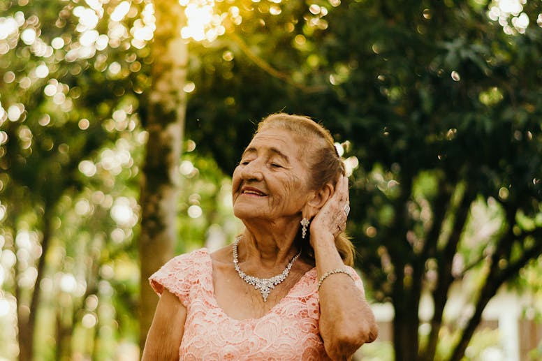 Graceful senior woman in a pink dress enjoying a peaceful moment outdoors amidst sunlit trees.