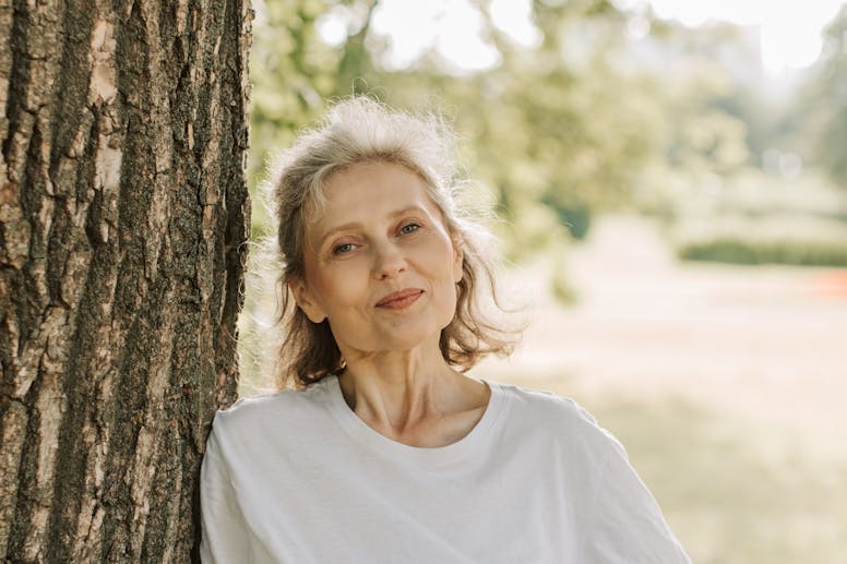 Senior woman leaning on tree with yoga mat and phone, enjoying a sunny day outdoors.