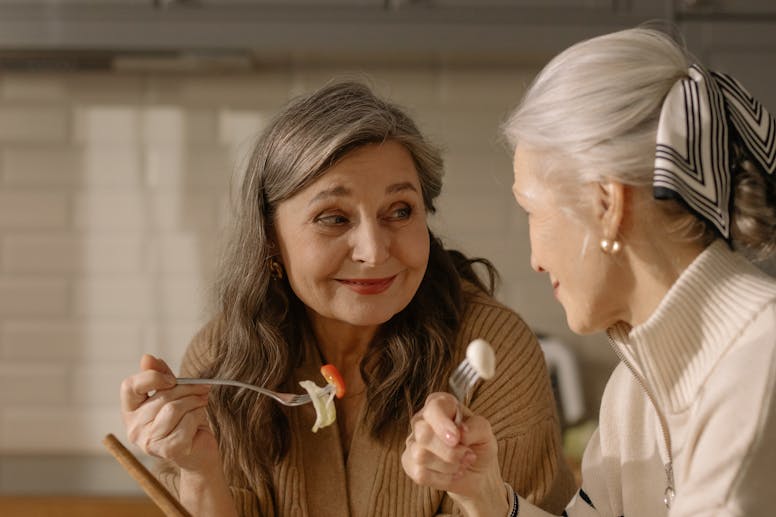 Two senior women share a light-hearted conversation over a fresh salad in a cozy kitchen.